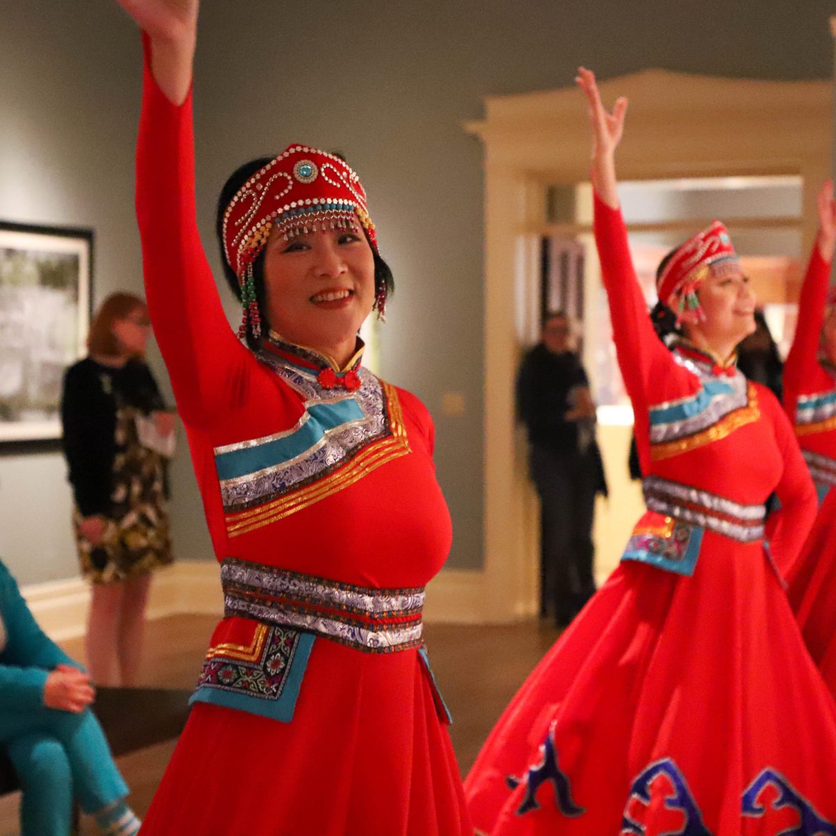 a line of female dancers in red dresses with Tibetan patterning, right arm raised in dance, performing for guests in gallery