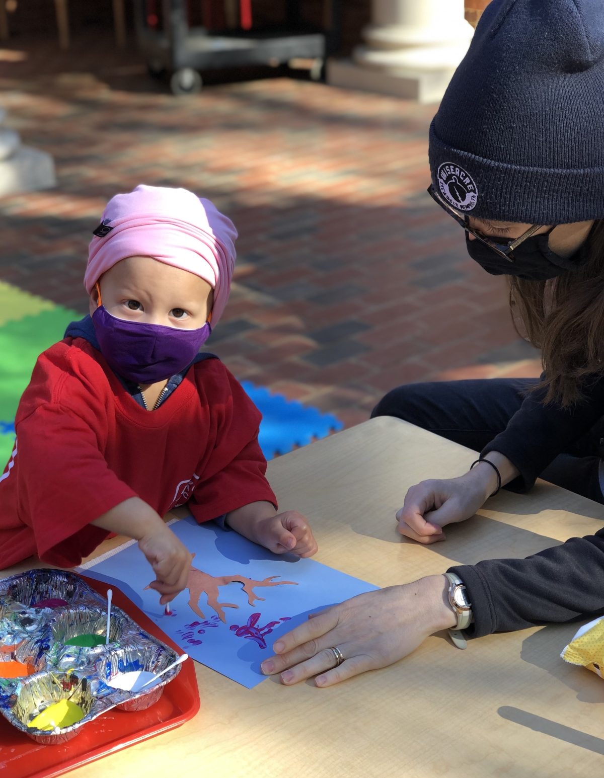 masked toddler pressing q-tip with purple paint onto paper tree, mom helping
