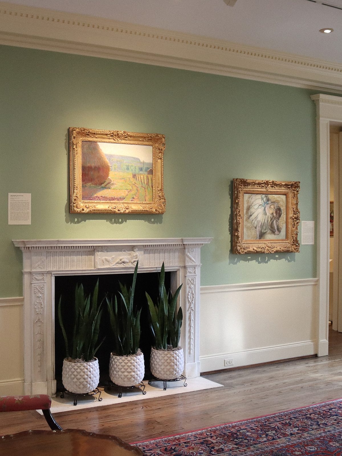 gallery with green walls, living room furniture, fireplace with three potted plants inside and three paintings hung above