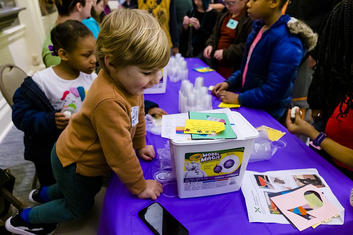 toddler looking at craft projects on table clothed in purple, surrounded by other kids