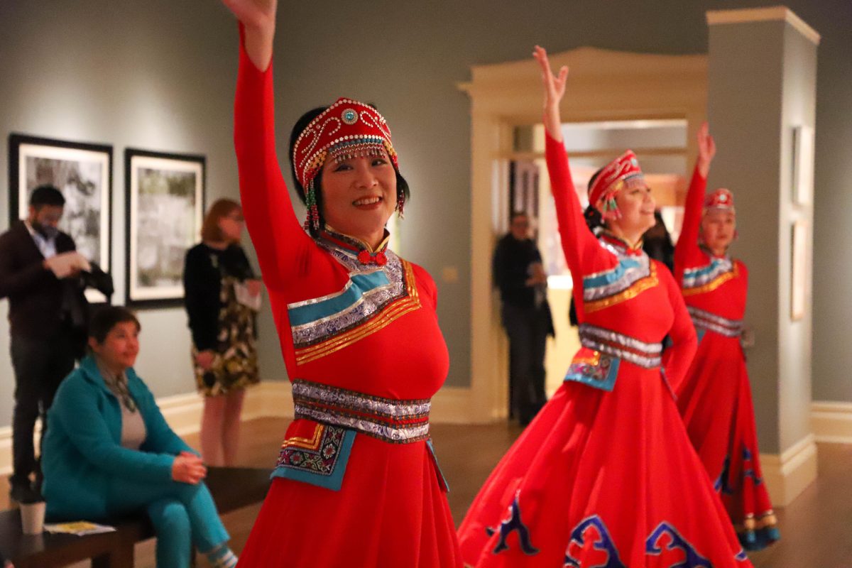 a line of female dancers in red dresses with Tibetan patterning, right arm raised in dance, performing for guests in gallery