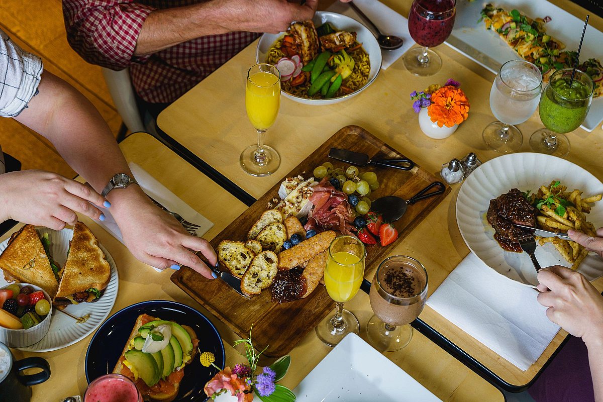 brunch meal laid out on table, three adults reaching for food on paper plates, overhead view