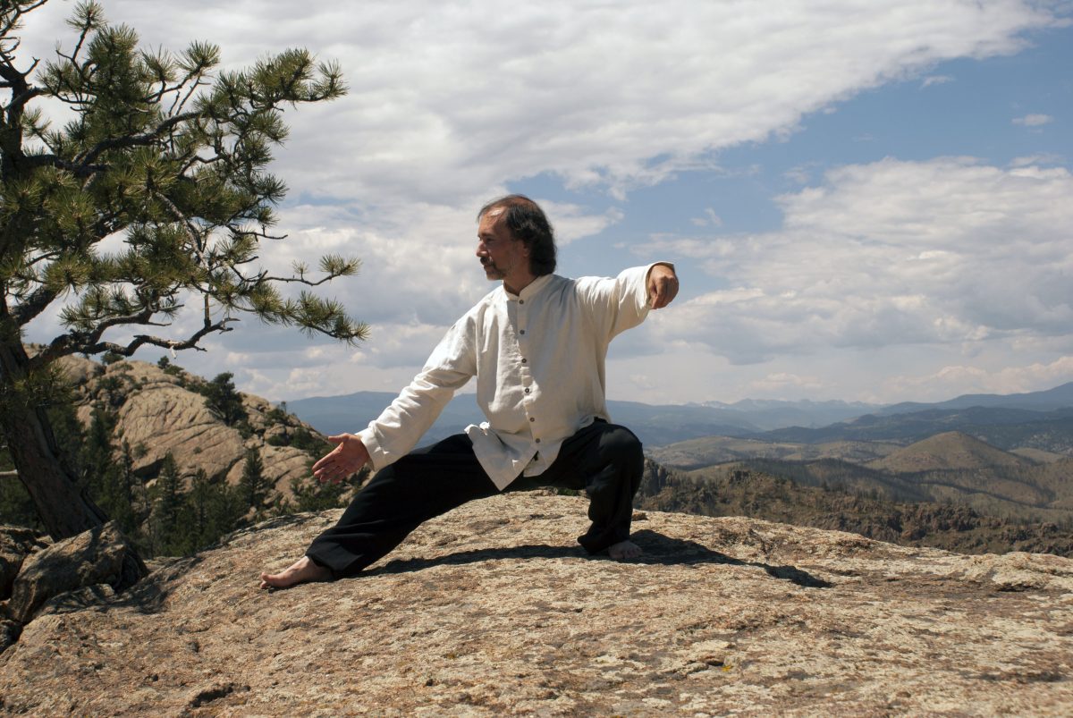 man in white and black in taijiquan pose atop mountain, hills and clouds in background