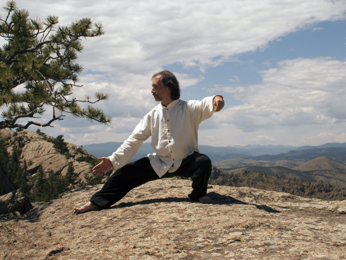 man in white and black in taijiquan pose atop mountain, hills and clouds in background