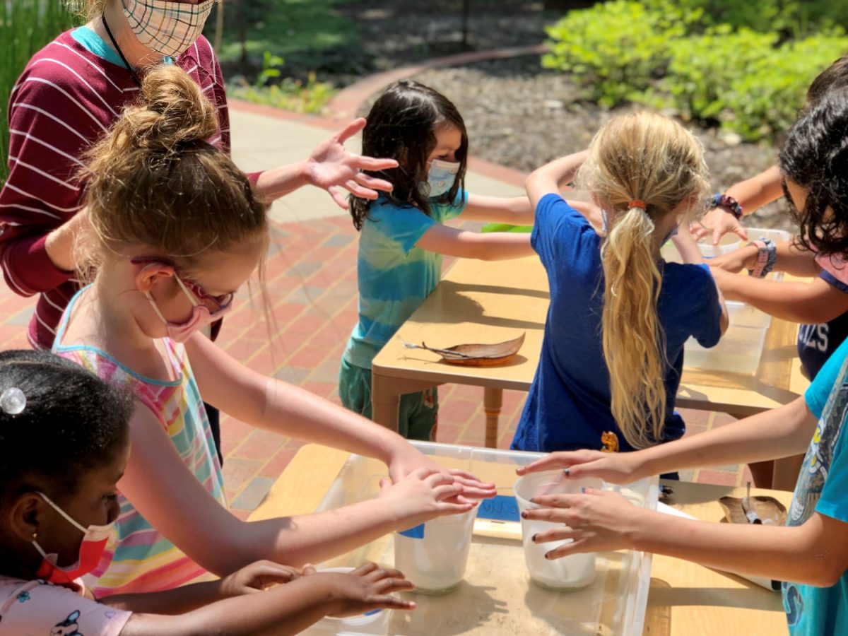 masked kids standing around a table outside, working on an art activity with instructor