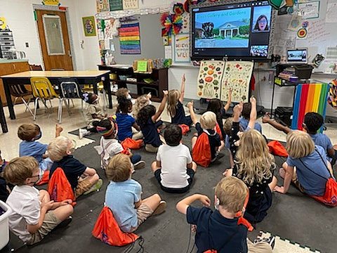 classroom of young kids sitting on floor with red drawstring backpacks watching a television zoom lesson from dixon employee