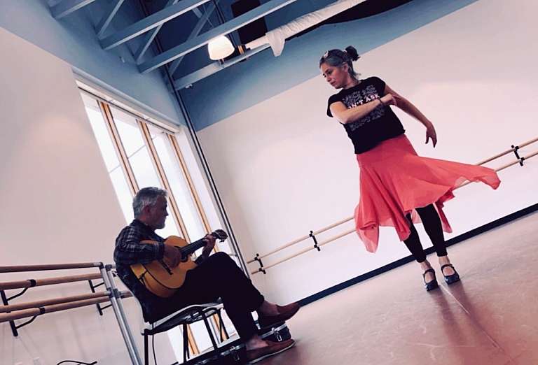 man in black sitting in chair playing guitar, woman in pink flamenco skirt dancing, both in studio